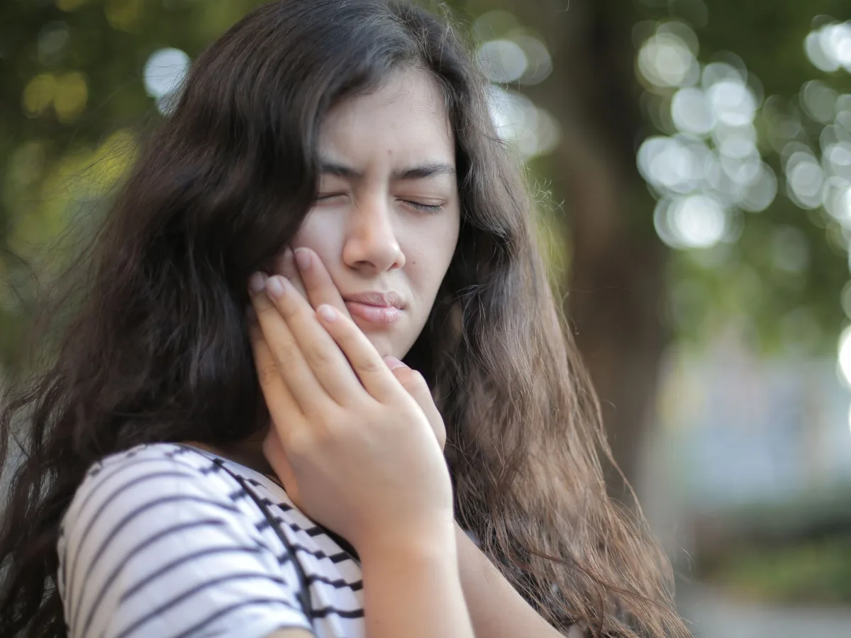 A young woman wincing with her eyes closed, pressing her fingers to the side of her jaw